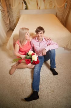 Couple sits on the floor of a hotel room, sharing a romantic moment with flowers, celebrating their love together.