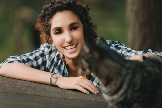 Curly-haired woman in a checkered shirt extends her arm to a tabby cat, smiling brightly. The cat appears slightly out of focus, emphasizing the woman's vivid, happy expression