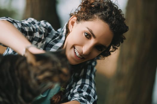 Woman with curly hair and checkered shirt joyfully reaches for a tabby cat. Her bright smile contrasts with the softly blurred background and the cat's slightly out-of-focus presence
