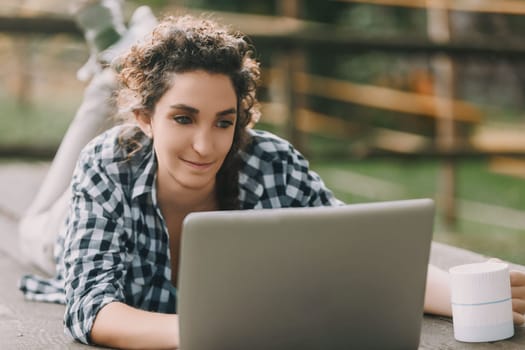 woman with curly braided hair and a checkered shirt lies on her stomach, working on a laptop. Her bright smile and nearby coffee mug suggest a happy, casual work environment