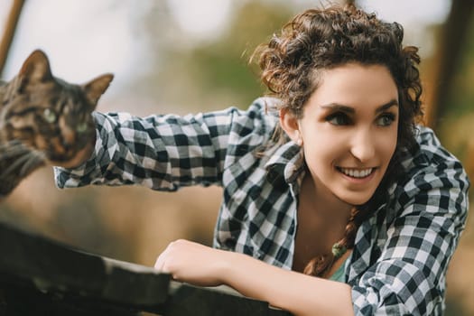 oung woman with curly hair and a checkered shirt smiles brightly while reaching out to pet a tabby cat. The cat is partially out of focus, highlighting the woman's joyful expression
