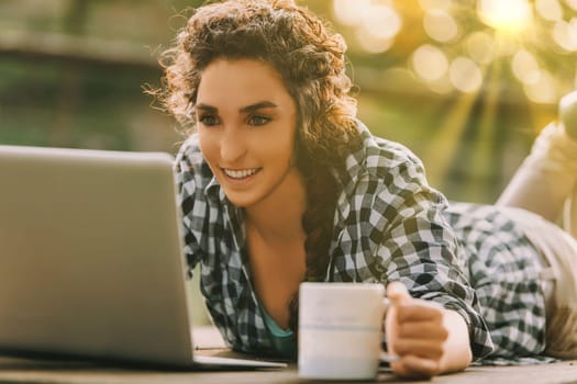 Curly-haired woman with a braid and checkered shirt lies on her stomach, typing on a laptop. Her cheerful smile and coffee mug convey a pleasant and engaging work environment
