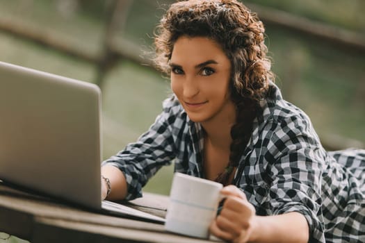 Young woman with curly hair in a braid, dressed in a checkered shirt, lies on her stomach using a laptop. Her broad smile and coffee mug create a relaxed and productive atmosphere