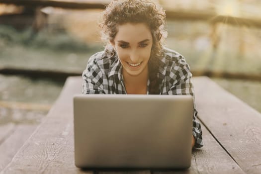 Young woman with curly hair in a braid, dressed in a checkered shirt, lies on her stomach using a laptop. She is deeply engrossed in her work, with the golden sunlight creating a cozy ambiance