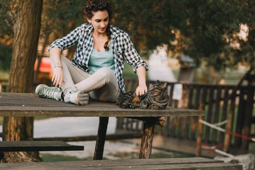 Woman with curly braided hair and a checkered shirt sits on a picnic table, petting a relaxed cat. Her casual pose and the serene outdoor setting highlight a peaceful moment with her pet