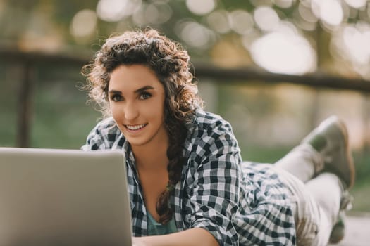 Woman with braided curly hair, wearing a checkered shirt, uses a laptop while lying on her stomach outdoors. Her concentrated expression and the warm sunlight create a serene working environment