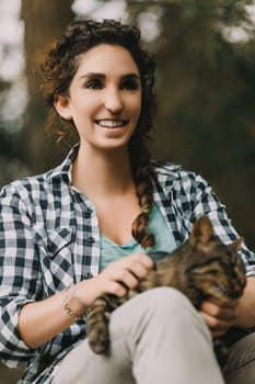 Smiling woman with curly hair and checkered shirt holds a blissful tabby cat, both enjoying the moment. The cat’s expression reflects pure contentment, as if experiencing a feline spa treatment