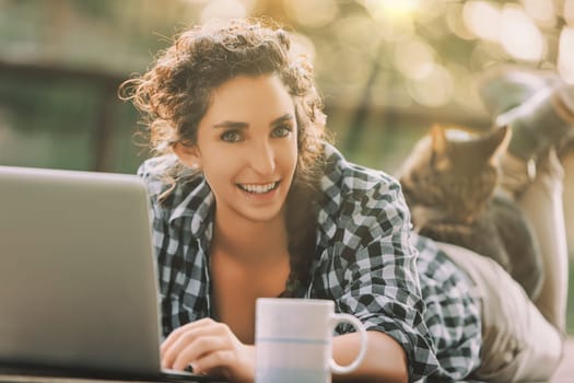 woman with curly braided hair and a checkered shirt lies on a bench, typing on a laptop. Her cat rests on her back, and a coffee mug is beside her, adding to the cozy scene