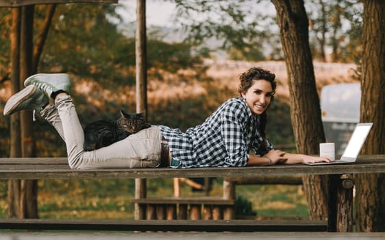 Woman with curly braided hair and a checkered shirt lies on a bench using a laptop. A cat rests on her back, adding a cozy touch to the outdoor work setting, with a coffee mug nearby