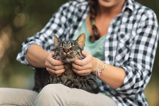 woman in a checkered shirt smiles, holding a delighted tabby cat. The cat’s relaxed demeanor suggests it is enjoying a spa-like pampering session