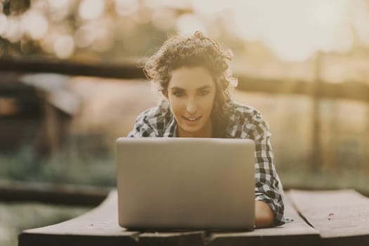 woman with curly braided hair and a checkered shirt lies on her stomach, focused on her laptop. The warm outdoor light and her intent expression highlight her engagement with her task