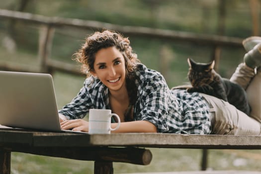 Woman with braided curly hair, wearing a checkered shirt, uses a laptop while lying on a bench. A cat naps on her back, and a coffee mug sits nearby, highlighting a serene outdoor setting