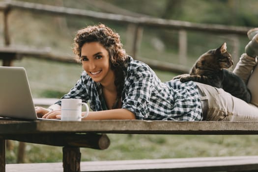 Curly-haired woman with a braid and checkered shirt lies on a bench, working on a laptop. Her cat rests on her back, and a coffee mug is beside her, creating a peaceful atmosphere