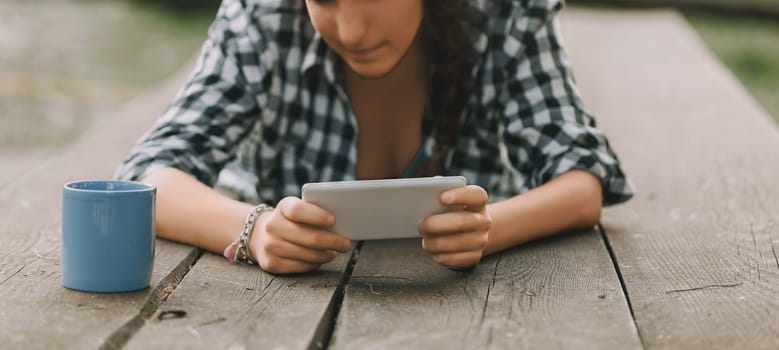 woman with curly braided hair and a checkered shirt lies on her stomach, engrossed in a smartphone. Her unrecognizable face and a blue mug rest on the wooden surface