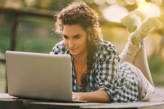 Curly-haired woman with a braid and checkered shirt lies on her stomach using a laptop. Her focused expression reflects deep engagement, complemented by the warm, golden outdoor light