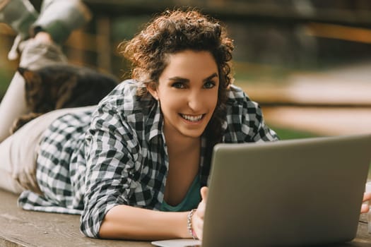 Young woman with curly hair in a braid, dressed in a checkered shirt, lies on a bench using a laptop. A cat sleeps on her back, creating a relaxed and comfortable atmosphere