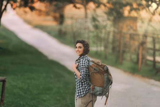 Woman with curly braided hair and a checkered shirt smiles while walking with a large backpack on a sunny path. Her cheerful expression and casual attire suggest she is enjoying a hike