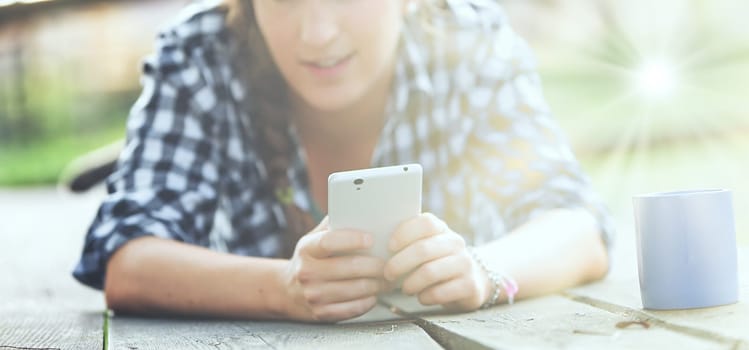 Woman with curly braided hair and a checkered shirt lies on her stomach outdoors, focusing on a smartphone. Her unrecognizable face and a blue mug create a peaceful, private scene