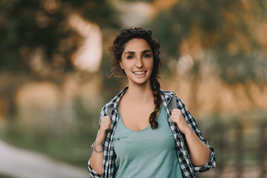 Young woman with curly braided hair and a checkered shirt smiles brightly while walking outdoors. Her joyful expression and relaxed posture highlight a serene and pleasant outdoor setting