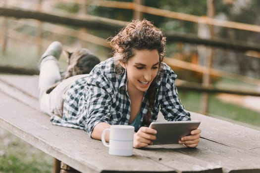 Young woman with curly hair in a braid, dressed in a checkered shirt and light pants, smiles while lying on a bench with a smartphone. A cat rests on her back, adding to the relaxed atmosphere