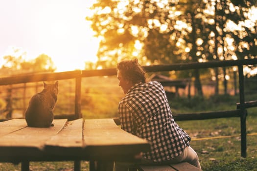 Woman with curly braided hair and a checkered shirt sits on a bench with her cat, both enjoying a serene sunset. Their relaxed postures reflect a peaceful retirement in a natural setting