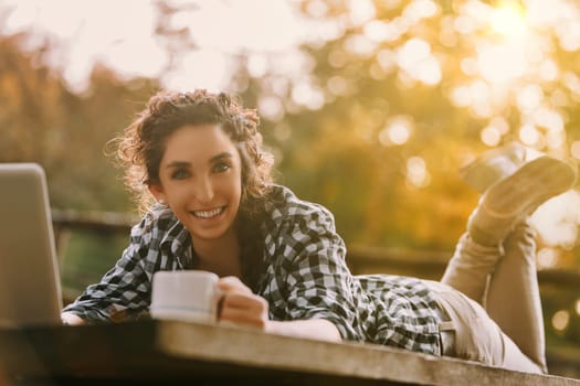 Woman with braided curly hair, wearing a checkered shirt, smiles broadly while using a laptop outdoors. A coffee mug next to her enhances the relaxed, productive setting