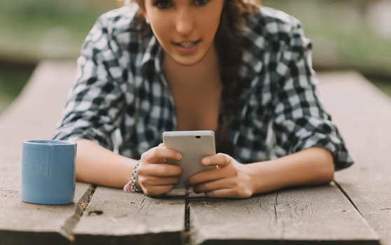 Young woman with curly hair in a braid, dressed in a checkered shirt, lies down while using a smartphone. Her unrecognizable face and a blue mug emphasize the serene setting