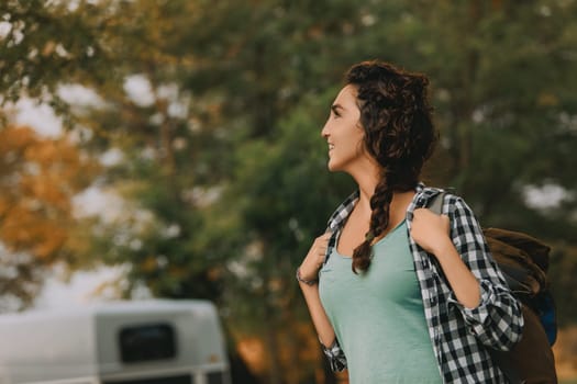 Curly-haired woman with a braid and checkered shirt smiles as she looks around, with a backpack on her shoulders. Her casual and adventurous spirit shines in the outdoor setting