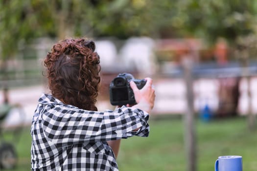 Woman with curly hair in a braid, wearing a checkered shirt, seen from behind as she holds a camera and takes a photo. The outdoor background is softly blurred, focusing on her actions