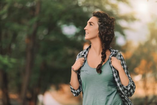 woman with curly braided hair and a checkered shirt smiles while looking around outdoors. Her backpack and casual attire suggest she is enjoying an adventurous day in nature