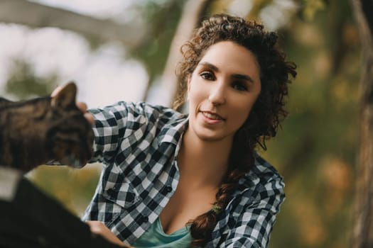 Smiling young woman in a checkered shirt reaches out to a tabby cat. Her curly hair and bright eyes are in sharp focus, while the cat remains slightly blurred in the foreground