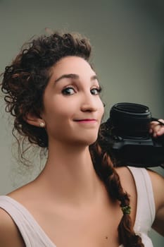 oung woman with curly hair and a braid, wearing a sleeveless top, holds a camera on her shoulder. She smiles subtly, exuding confidence and readiness for a photography session