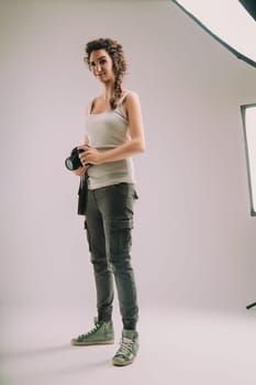 Confident woman with curly hair in a braid, wearing a sleeveless top and cargo pants, holds a camera. She stands in a studio, exuding a strong sense of pride and enthusiasm for her photography role