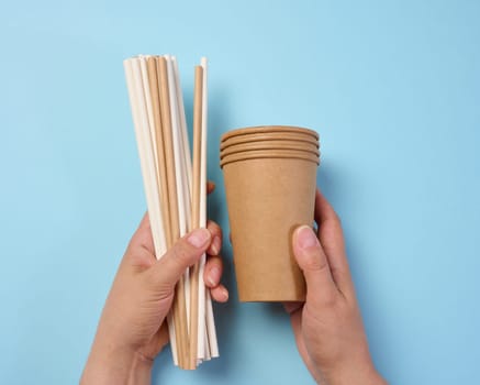 Female hand holding paper cocktail straws and paper cups on a blue background, top view