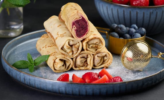 Crepes stacked in a pile, one of which is cut and filled with berry filling. The plate decorated with blueberries, black background