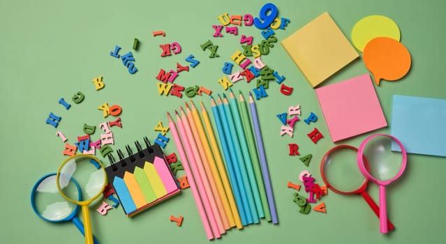 Stationery laid out on a green background. Multi-colored pencils ,  sticky notes and wooden letters, preparing for study or work, organizing workspace