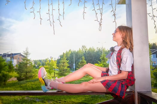 A girl relaxes on a wooden railing in a sunny neighborhood, wearing headphones and looking content as she enjoys the view.