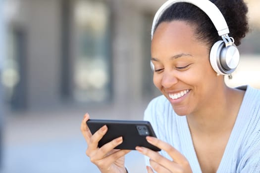 Happy black woman wearing headphone watching videos on phone in the street