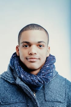 Young man with a confident expression wearing a blue patterned scarf and a grey wool coat. His short hair is neatly trimmed, and he has a small earring in his left ear
