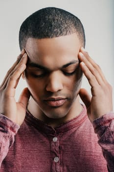 Young man with a pained expression, pressing his fingers to his temples. Wearing a maroon shirt, he appears to be experiencing a headache or intense stress, reflecting discomfort and frustration