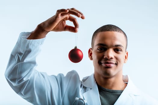 Doctor in a white lab coat, smiling and holding a red Christmas ornament. As a military doctor, he seeks to enhance his patients' health during the festive Christmas period