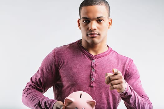 Young man in a maroon henley shirt, holding a piggy bank and pointing forward. His serious expression suggests advising caution with savings and the importance of investing wisely to avoid losses
