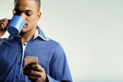 Individual with a serene look, holding a blue coffee mug and a smartphone. Dressed in a blue shirt, he seems to be enjoying a peaceful moment and staying informed