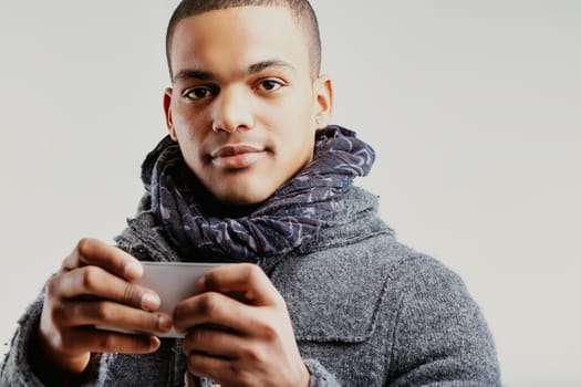 oung man, dressed in a grey coat and scarf, smiling as he holds a smartphone. His warm expression suggests he is engaged in a positive and enjoyable activity