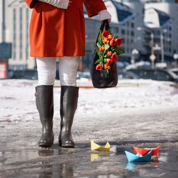 Closeup of woman in a orange coat and rubber boots walking down the street with a bouquet of tulips in spring city