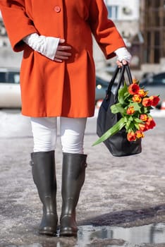 Closeup of woman in a orange coat and rubber boots walking down the street with a bouquet of tulips in spring city