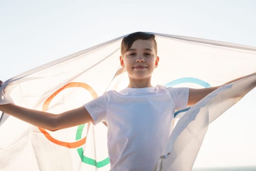 BARNAUL CITY. RUSSIA - JUNE 08, 2021: Portrait of boy waving flag the Olympic Games outdoors over cloudy sunset sky. Children sports fan. Summer olympic games concept on June 08, 2021 in Altayskiy krai, Siberia, Barnaul, Russia