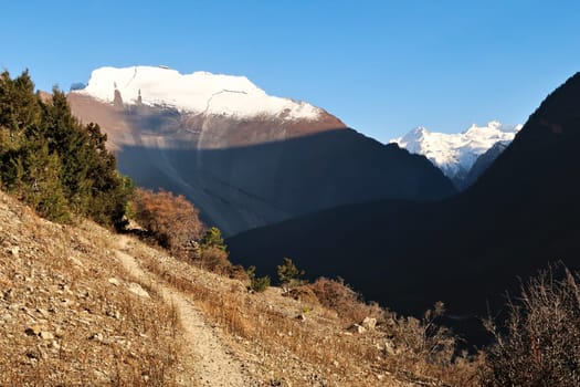A small trail in the village of Upper Pisang, the smooth western rock face of Paungda Danda, the Great Wall of Pisang and the peak of Lamjung Kailas in the background, Annapurna Circuit Trek, Nepal 2022