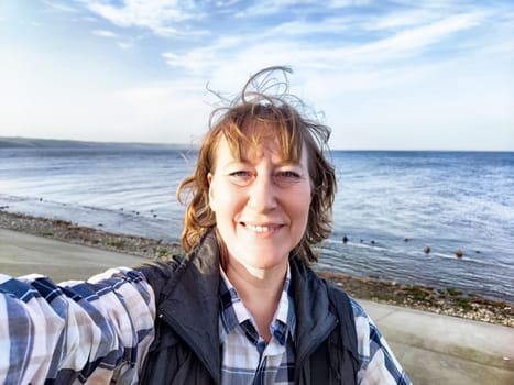 A middle-aged blogger enjoys a sunny beach day, taking a joyful selfie with the ocean behind her.