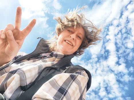 A middle-aged female travel blogger smiles happily while capturing a selfie outdoors under a clear blue sky with clouds.